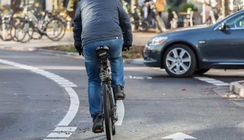 Un cycliste vu de dos roule sur une piste cyclable, avec une voiture grise garée à côté.