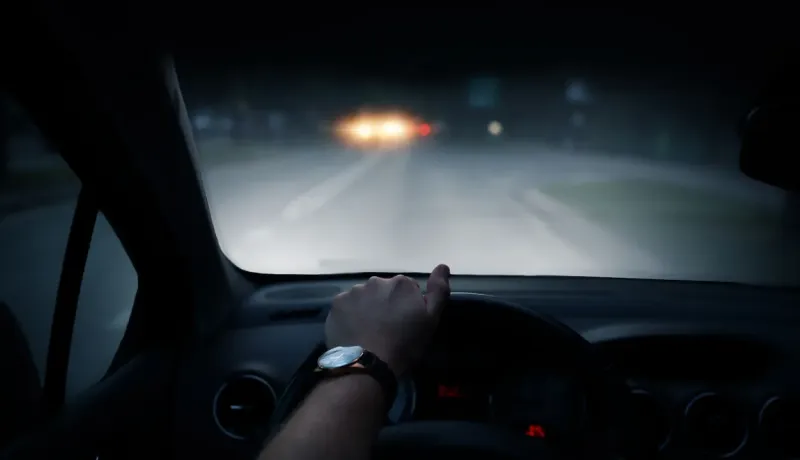 Une main tient le volant d'une voiture, les phares d'un autre véhicule sont visibles de nuit à travers le pare-brise.