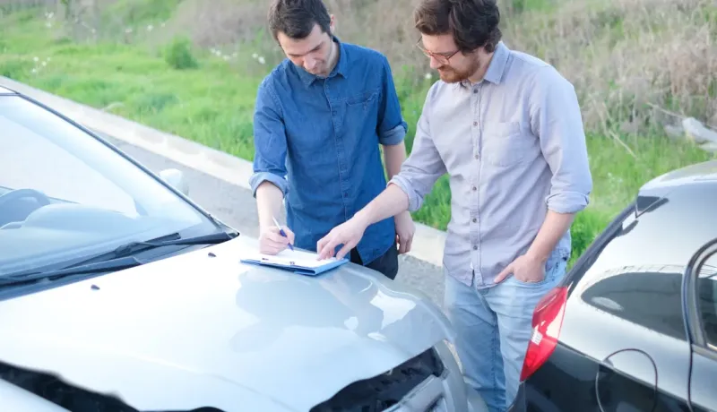 Deux hommes sont penchés sur le capot d'une voiture endommagée, l'un d'eux remplit un constat à l'amiable sur un presse-papiers.