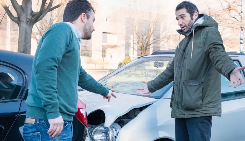Deux hommes discutent avec des gestes expressifs devant les capots endommagés de leurs voitures après un accident.