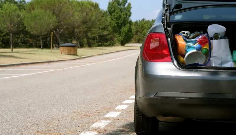 Le coffre ouvert d'une voiture grise révèle des sacs et des objets, garée sur un parking ensoleillé.