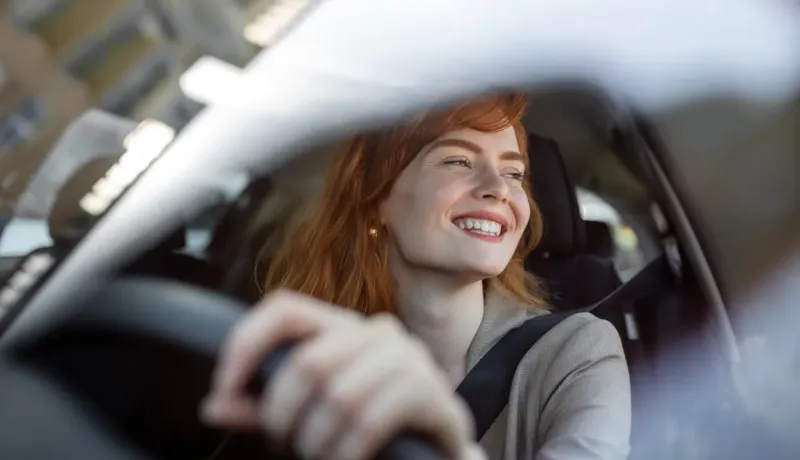 Une femme rousse souriante et joyeuse est vue à travers le pare-brise, conduisant sa voiture.
