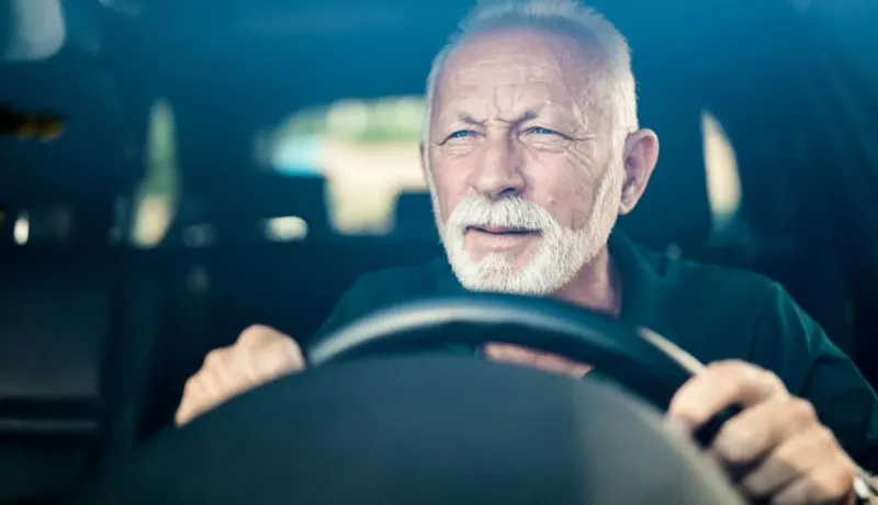 Un homme âgé aux cheveux et à la barbe blancs conduit une voiture, le regard concentré.