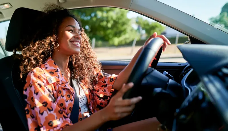 Une jeune femme aux cheveux bouclés sourit en conduisant sa voiture par une journée ensoleillée.