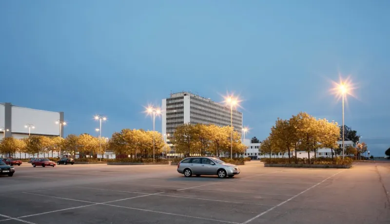 Un parking presque vide, une voiture grise est au milieu de l'iamge et un bâtiment moderne en arrière-plan, éclairé par des lampadaires.