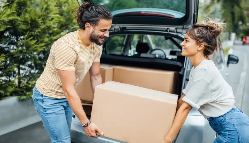 Un jeune couple charge des cartons dans le coffre ouvert d'une voiture, souriant l'un à l'autre.