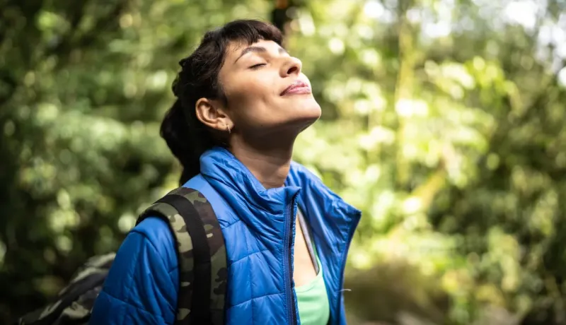 Une femme ferme les yeux et respire profondément dans une forêt ensoleillée.