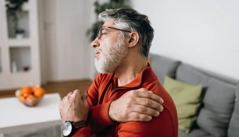 Un homme barbu aux lunettes, le visage grimaçant de douleur, se tient l'épaule.