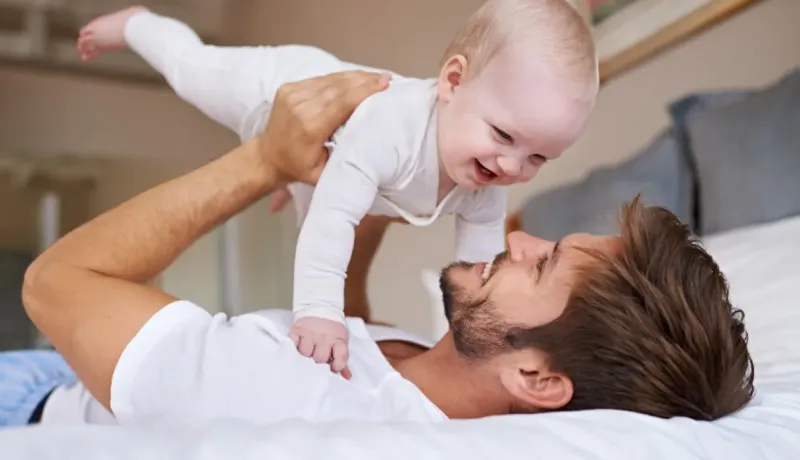 Un père heureux joue avec son bébé, le tenant au-dessus de lui sur le lit.