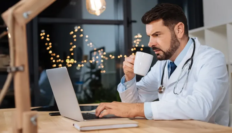 Un médecin en blouse blanche boit du café en travaillant sur son ordinateur portable de nuit.