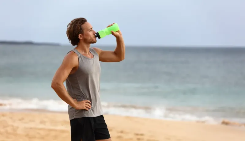 Un homme boit de l'eau après l'exercice sur une plage, en regardant l'océan.