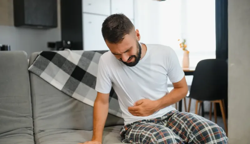 Un homme barbu assis sur un canapé, se tenant le ventre, exprimant une douleur abdominale.