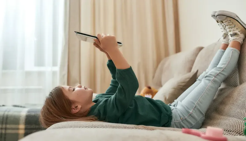 Une petite fille est allongée sur un canapé, les pieds en l'air, regardant une tablette.