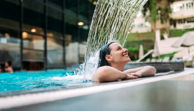 Une femme souriante se détend sous une cascade d'eau dans une piscine de spa.