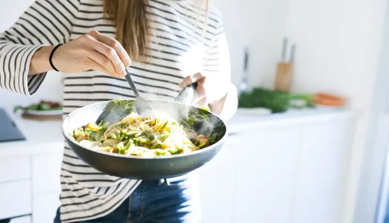 Gros plan sur une personne remuant des légumes et des pâtes dans une poêle dans une cuisine lumineuse.