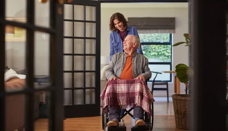 Une femme aide un homme âgé en fauteuil roulant à travers une porte ouverte.