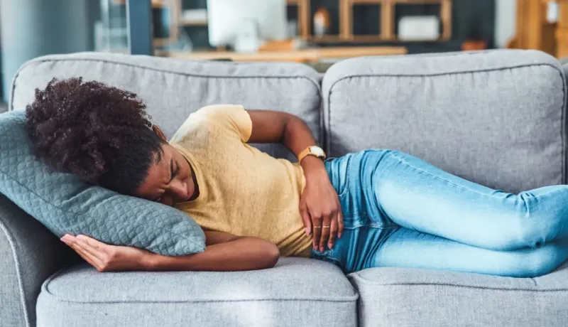 Une femme est allongée sur un canapé, les mains sur le ventre, exprimant une douleur.