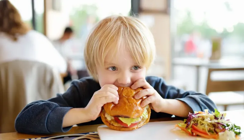 Un jeune garçon aux cheveux blonds mord dans un grand hamburger avec un regard espiègle.