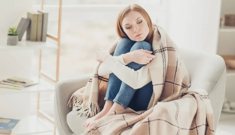 Une jeune femme triste, emmitouflée dans une couverture, assise seule sur un fauteuil.