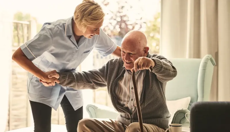 Une infirmière souriante aide un homme âgé à se lever de son fauteuil avec une canne.