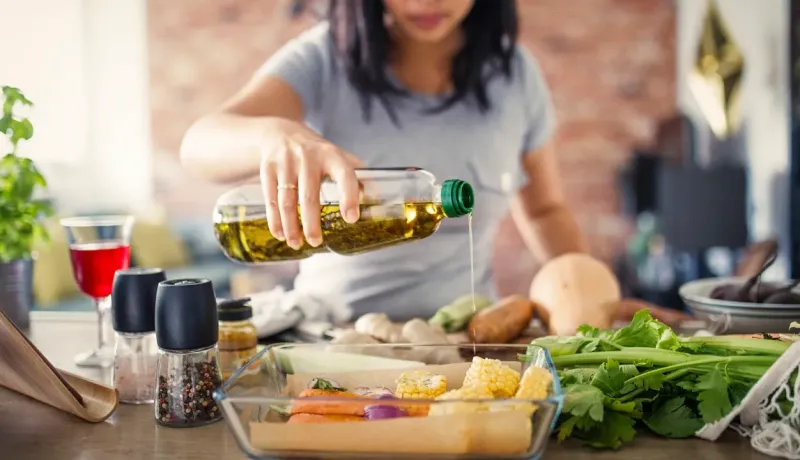 Une femme verse de l'huile d'olive sur des légumes dans un plat en verre, préparant un repas sain.