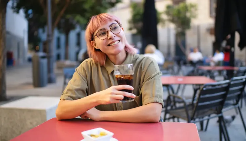 Une jeune femme aux cheveux roses et lunettes souriante, sirotant une boisson sur une terrasse de café.