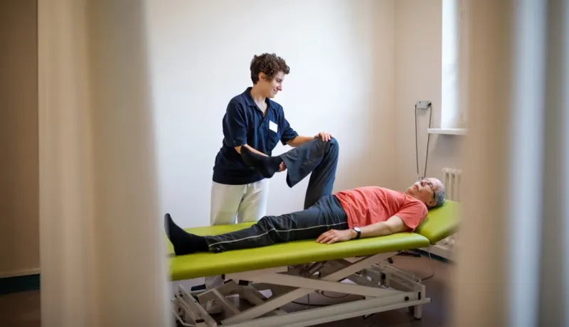 Un physiothérapeute aide un patient âgé à faire des exercices de jambe sur une table de rééducation.