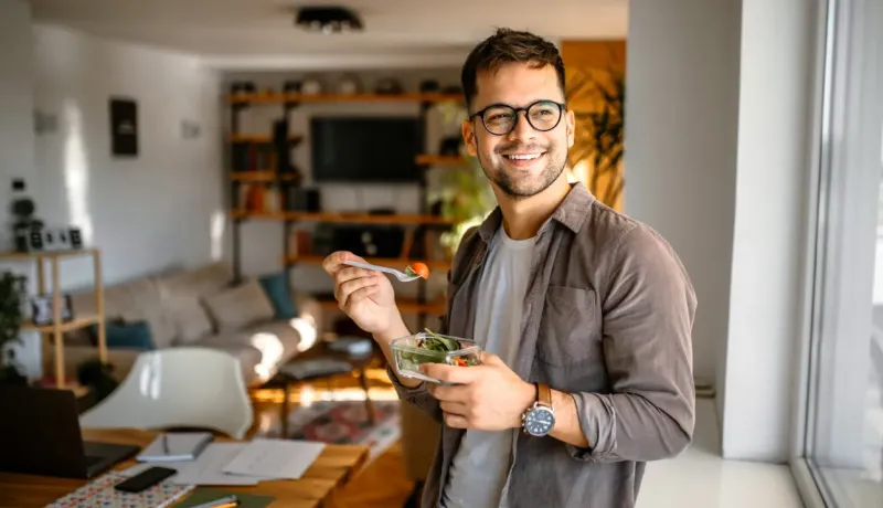 Un jeune homme souriant et lunettes mange une salade fraîche dans un récipient dans un intérieur moderne.