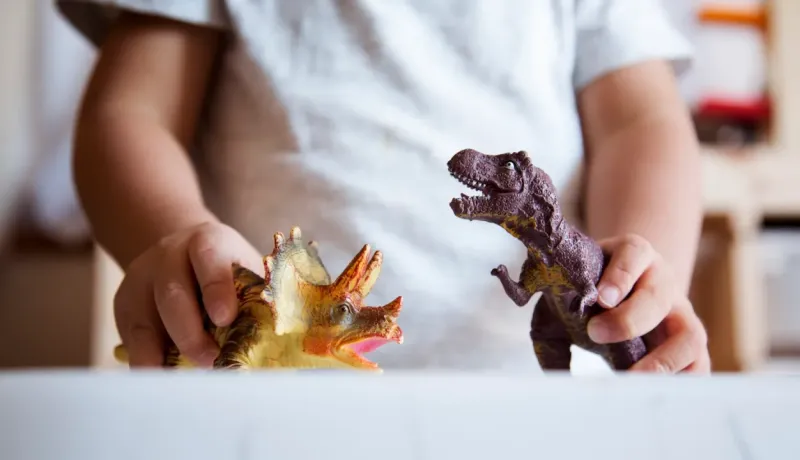 Gros plan sur les mains d'un enfant jouant avec deux figurines de dinosaures sur une table.