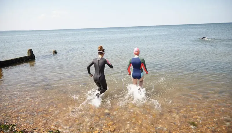 Deux nageuses en combinaison s'élancent dans la mer depuis une plage de galets par temps ensoleillé.