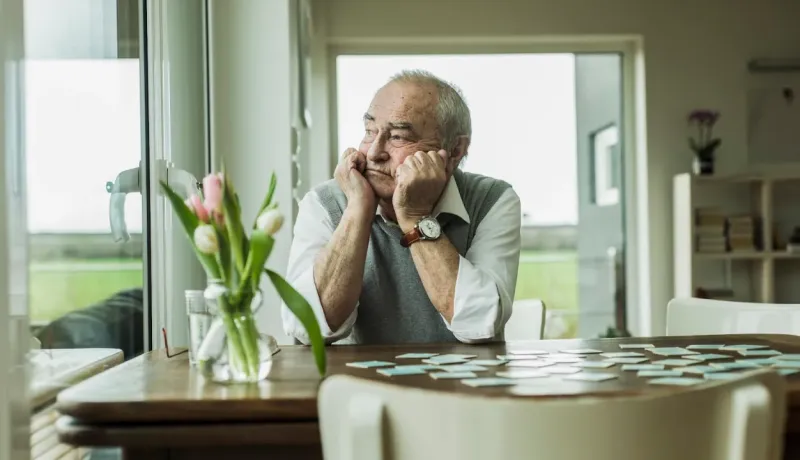 Un homme âgé pensif est assis à une table, des cartes éparpillées devant lui. Des fleurs sont dans un vase sur la gauche.