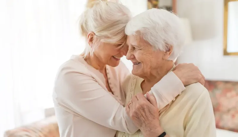 Deux femmes âgées se serrent dans les bras et sourient, un lien affectueux transparaît entre elles.