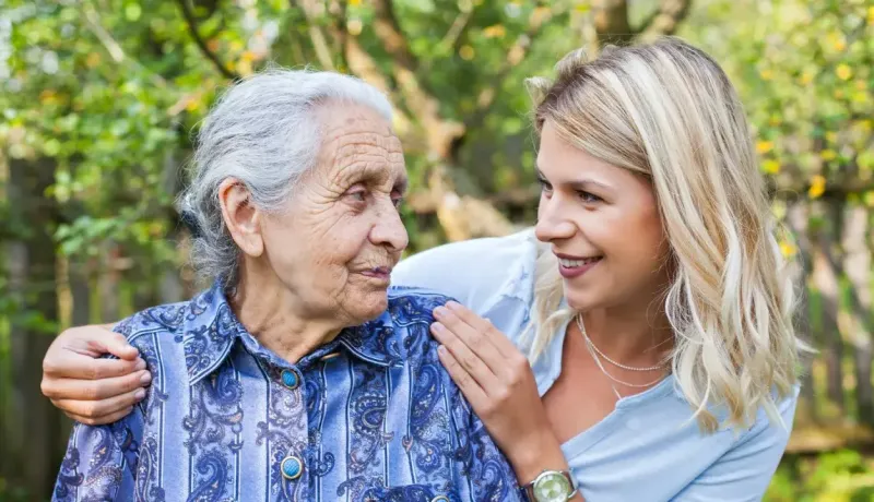 Une jeune femme souriante enlace une femme âgée en extérieur, partageant un moment de tendresse intergénérationnelle.