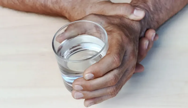 Gros plan sur les mains d'un homme âgé tenant fermement un verre d'eau clair sur une surface en bois.