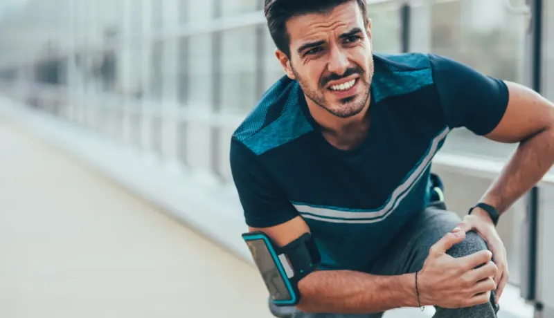 Un homme en tenue de sport grimace et se tient le genou, exprimant une douleur intense pendant l'effort.