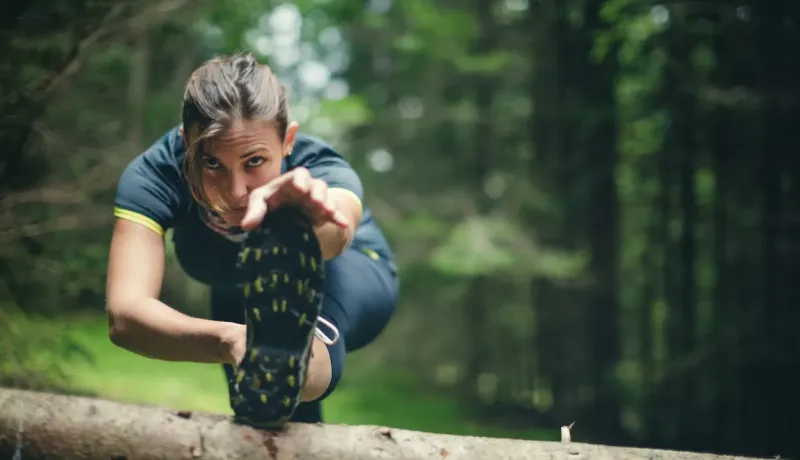 Une femme en tenue de sport s'étire en forêt, posant son pied sur une bûche, se concentrant sur l'exercice.