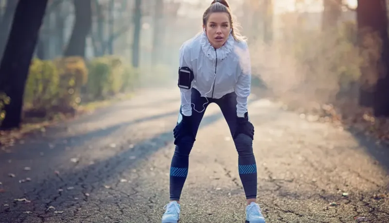 Une coureuse s'arrête pour reprendre son souffle sur un chemin, penchée en avant, les mains sur les genoux, par temps froid.