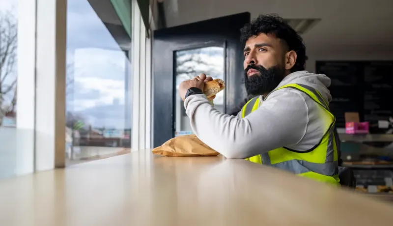 Un homme barbu en gilet de sécurité mange un sandwich près d'une fenêtre, regardant au loin.