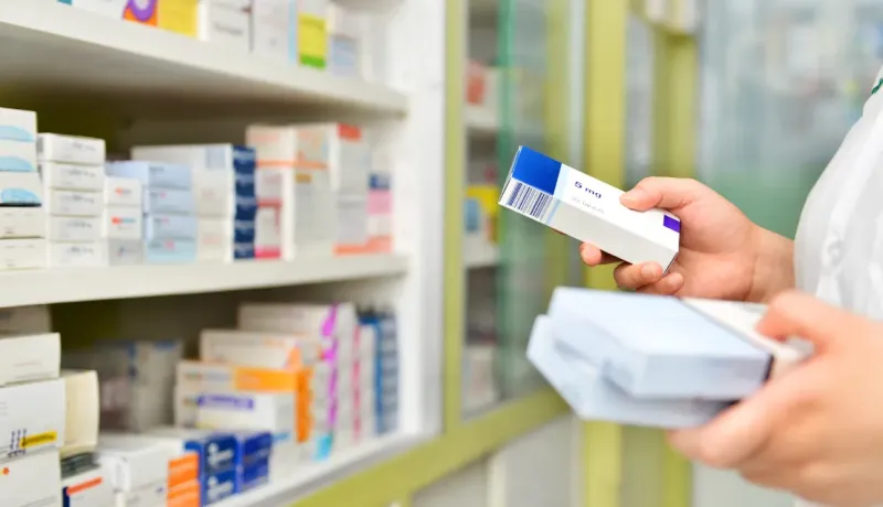 Un pharmacien prend une boîte de médicaments sur une étagère, entouré de nombreuses boîtes empilées.