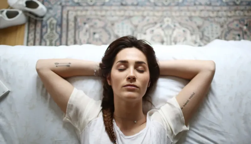 Une jeune femme allongée sur le dos, les mains derrière la tête, les yeux fermés, se détend sur un lit.