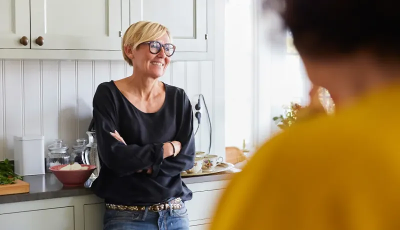 Une femme souriante aux cheveux courts et portant des lunettes, les bras croisés, discute dans une cuisine lumineuse.