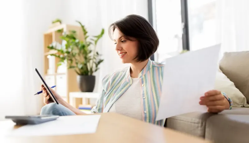 Une femme souriante assise sur le sol chez elle, tenant une tablette et des documents, gérant ses papiers.