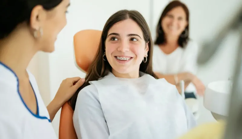 Une adolescente souriante avec un appareil dentaire est assise dans un fauteuil de dentiste, discutant avec le personnel.
