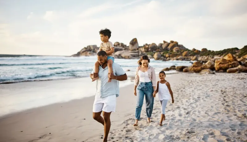 Une famille marche sur une plage de sable fin, le père portant son jeune fils sur les épaules, profitant du bord de mer.