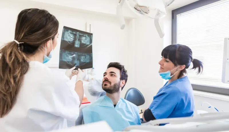 Deux dentistes examinent une radiographie dentaire avec un patient assis sur le fauteuil, expliquant les résultats.