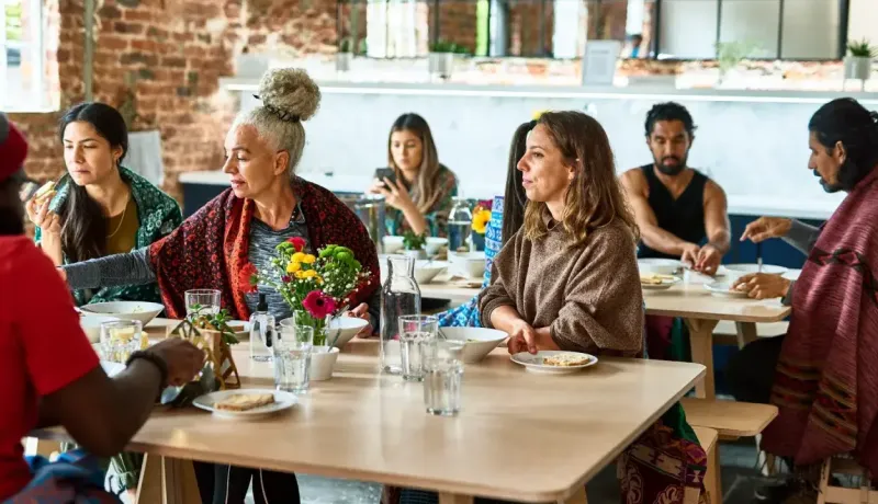 Un groupe de personnes diverses partage un repas à de grandes tables, engageant la conversation.