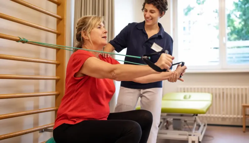 Un physiothérapeute aide une patiente à faire un exercice de rééducation avec une bande élastique contre un espalier.