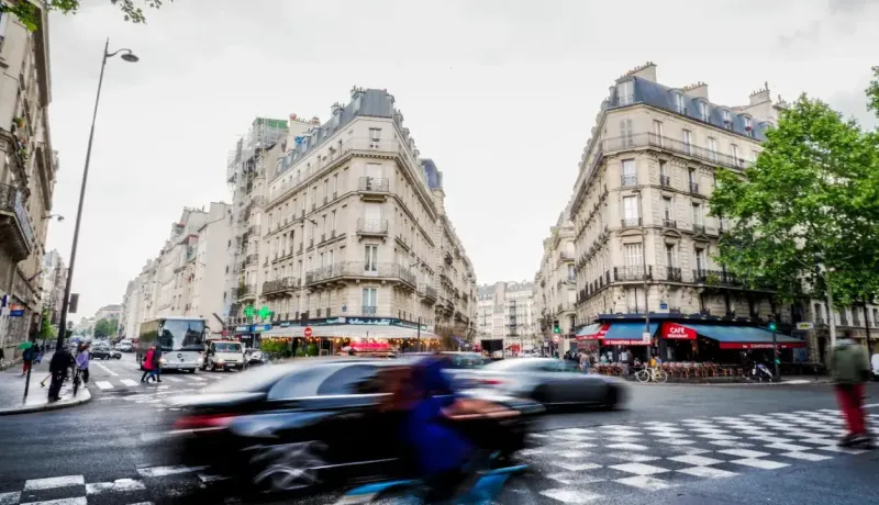 Scène de rue animée à Paris avec circulation et passants, bâtiments haussmanniens caractéristiques en arrière-plan.
