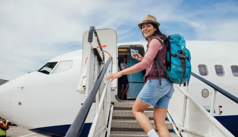 Une jeune femme souriante avec un sac à dos monte l'escalier d'un avion, prête pour le voyage.