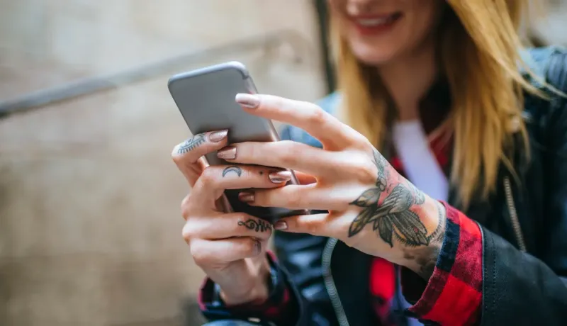 Gros plan sur les mains tatouées d'une jeune femme utilisant un smartphone, son visage flou en arrière-plan.